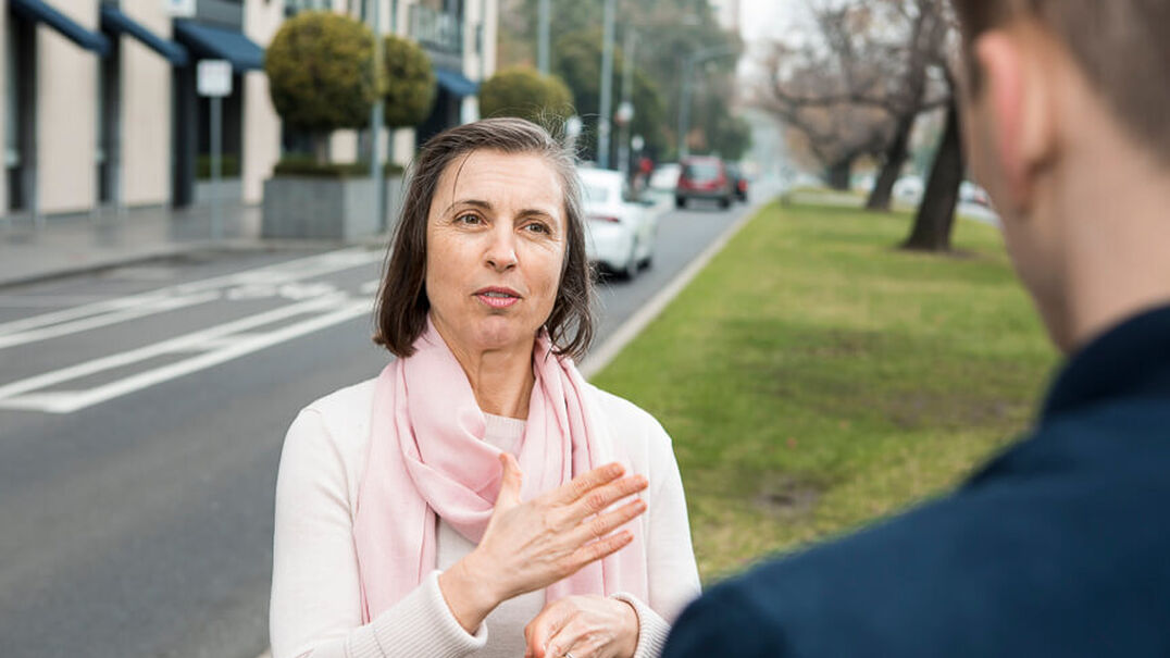 A woman wearing a pink scarf is having a conversation with a man using Auslan. The are standing on a grassy area next to a city street. Learn Auslan with Expression Australia.