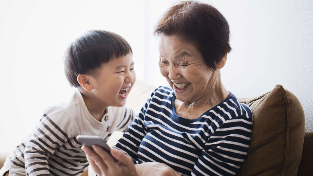 A grandmother a her toddler grandson are laughing while looking at a smart phone. If your hearing challenges are impacting your relationships Expression Audiology can help.