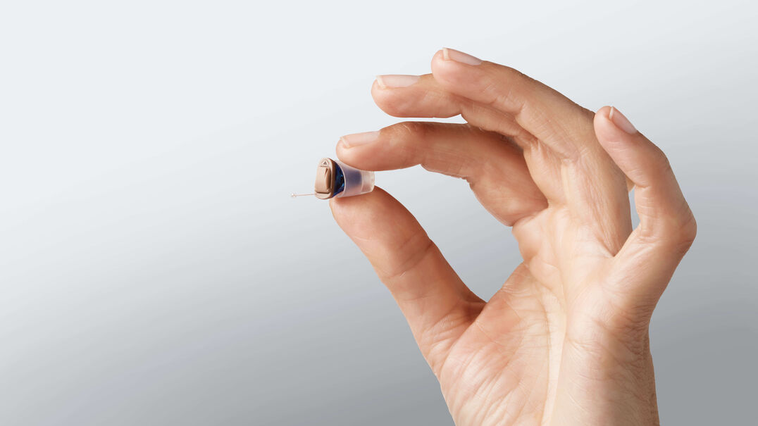 A hand holds a tiny in-the-ear hearing aid between its thumb and index finger. Expression Audiology can help you find the right hearing aid to suit your lifestyle.