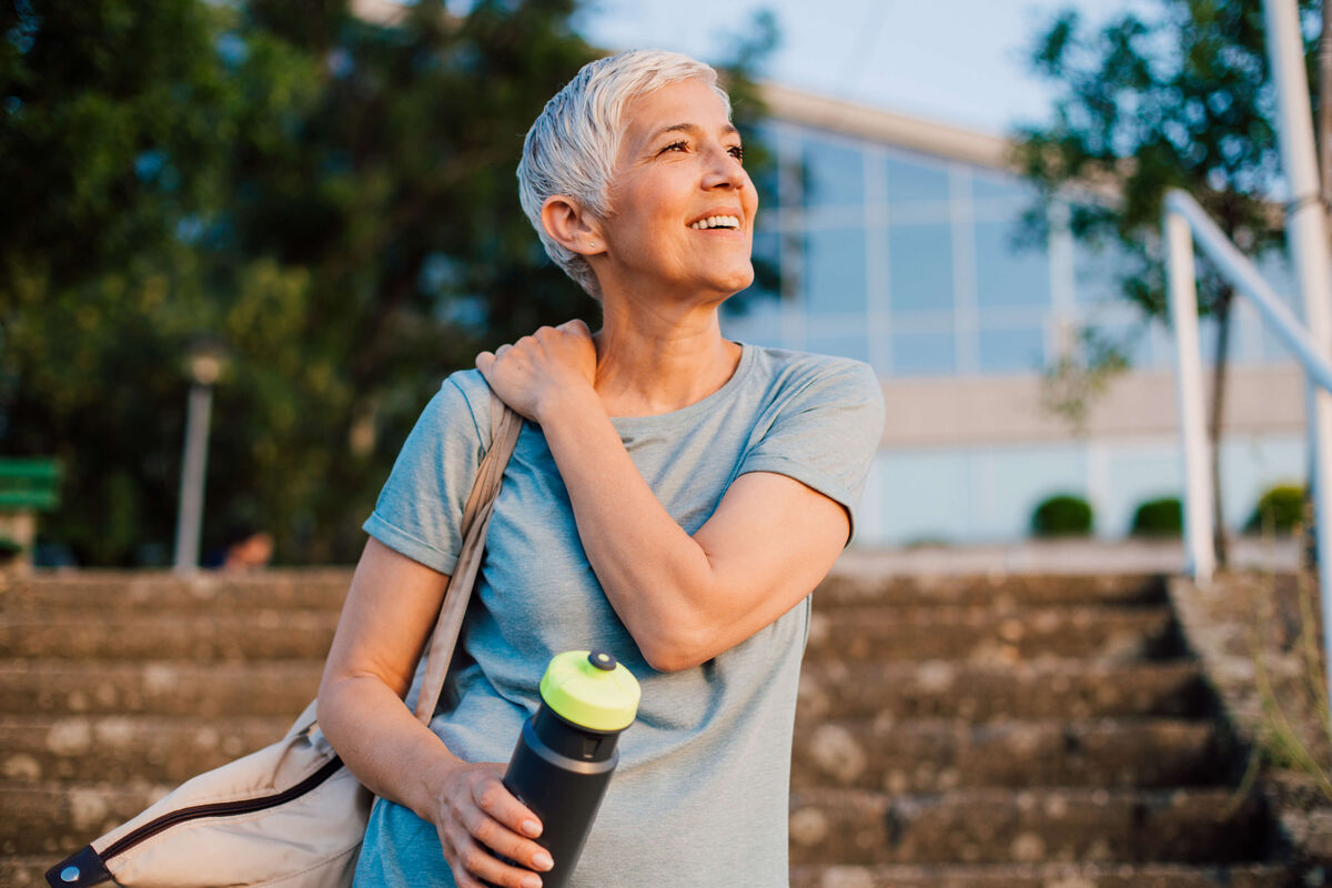 A woman smiles as she is bathed in golden sunshine. She is carrying a water bottle, suggesting she has been exercising. Don't let tinnitus cast a shadow over your life, talk to someone who understands at Expression Audiology.