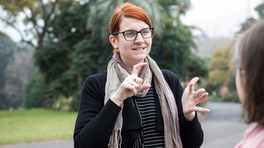 A woman with red hair and glasses is having a conversation using Auslan with another person in a park. Expression Australia Auslan courses are perfect for beginners.