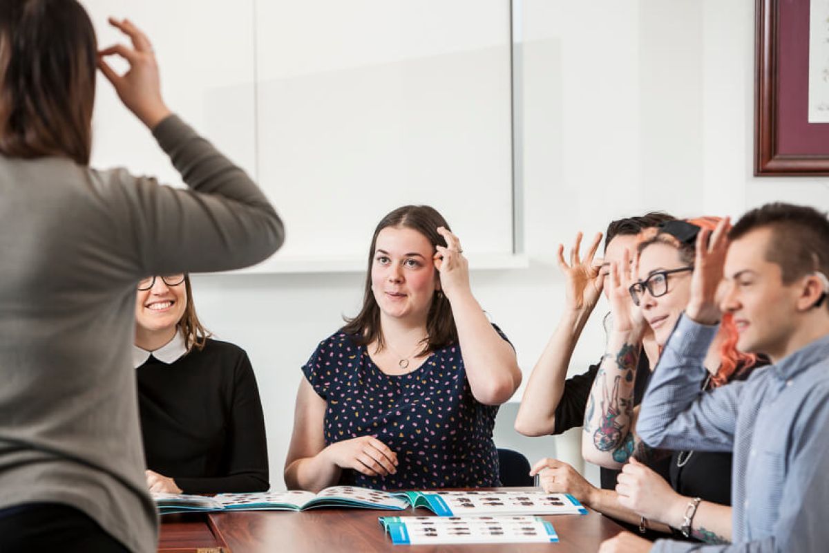 A class of Auslan students sitting at a table are watching their Auslan teacher sign and are mimicking the sign. Learn Auslan with Expression Australia.
