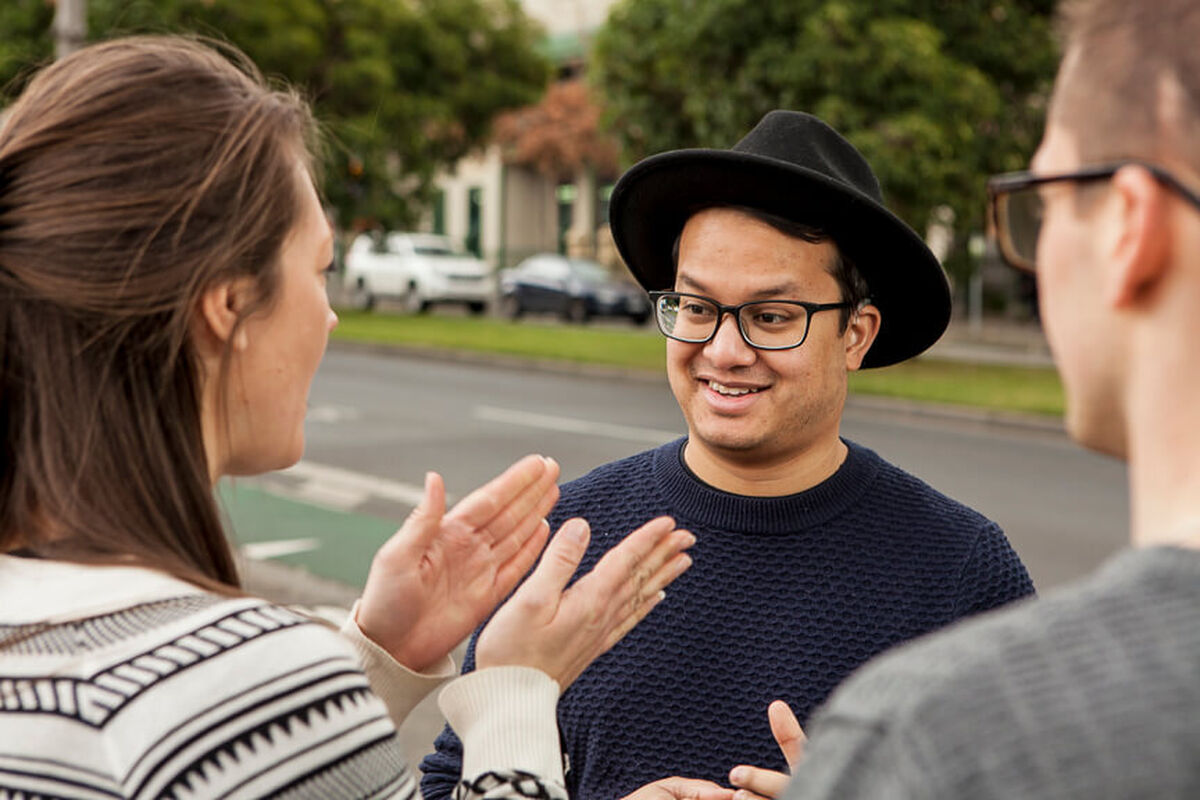 Two young men and a young woman are having a conversation in Auslan. One of the young men who is wearing a fashionable wide-brim hat is looking amused at what the woman is signing.
